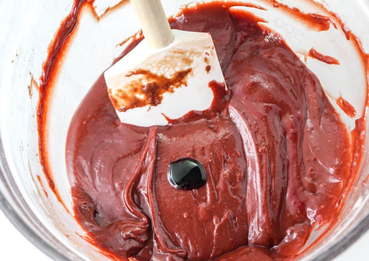 red food coloring being added to the batter in a glass mixing bowl