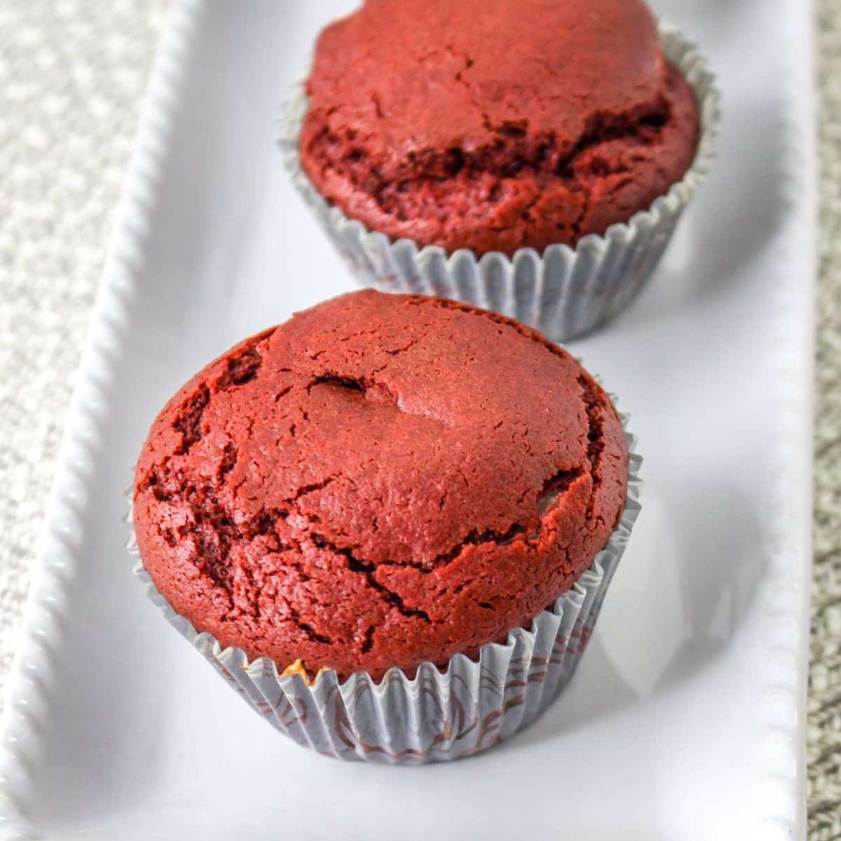 Two red velvet muffins, made using a classic red velvet muffins recipe, are placed in silver cupcake liners on a rectangular white ceramic tray.