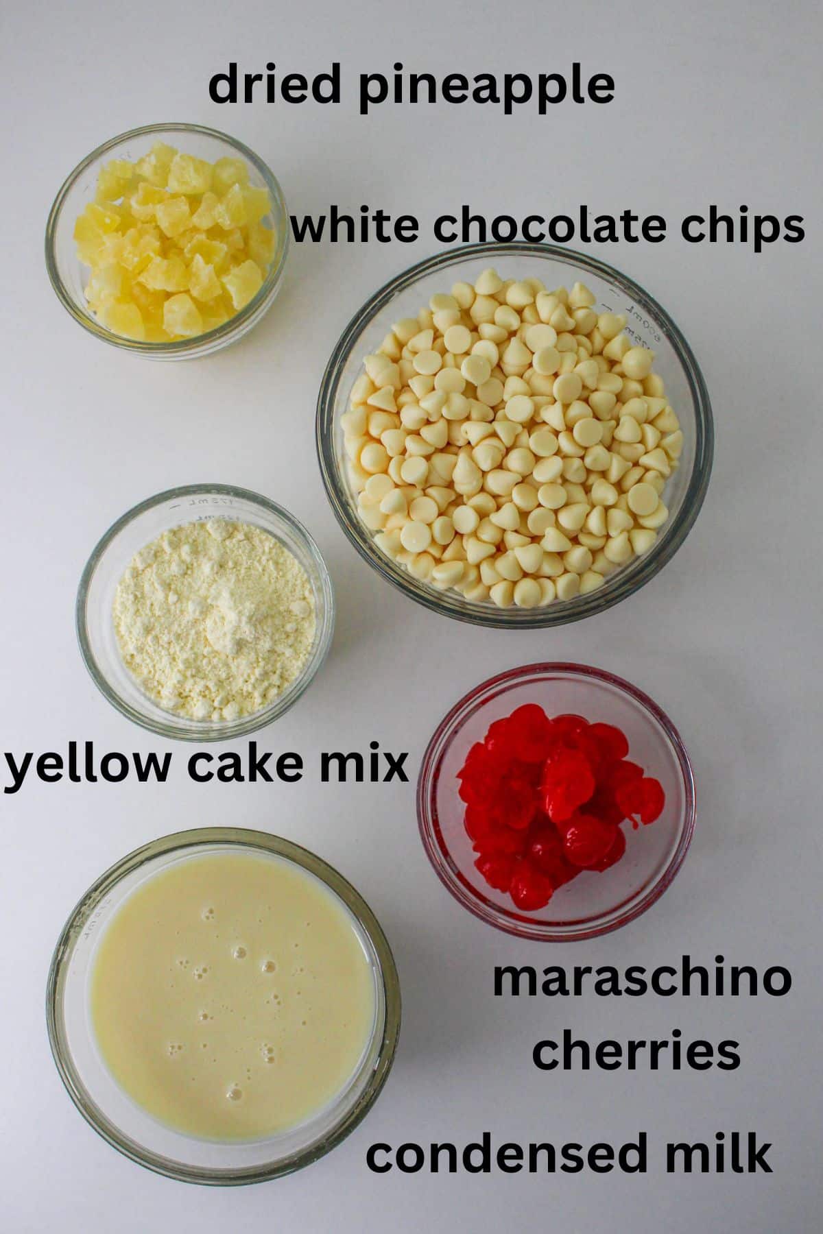 bowls of dried pineapple, white chocolate chips, yellow cake mix, maraschino cherries and condensed milk on a white background