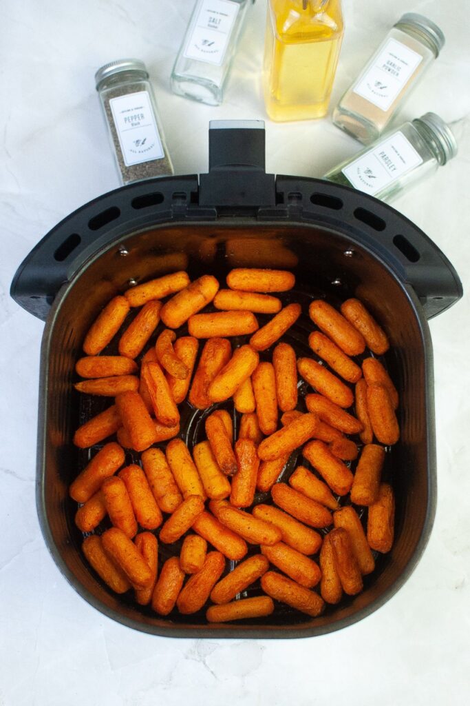 cooked baby carrots in an air fryer basket with gars of seasoning in the background