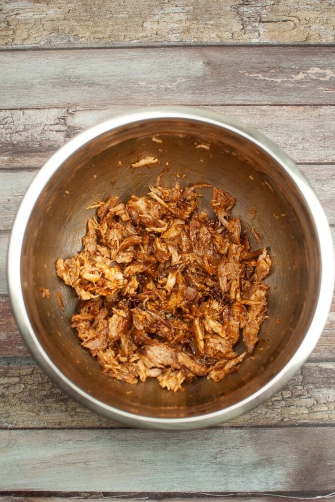 shredded pork topped with bbq sauce in a stainless steel mixing bowl