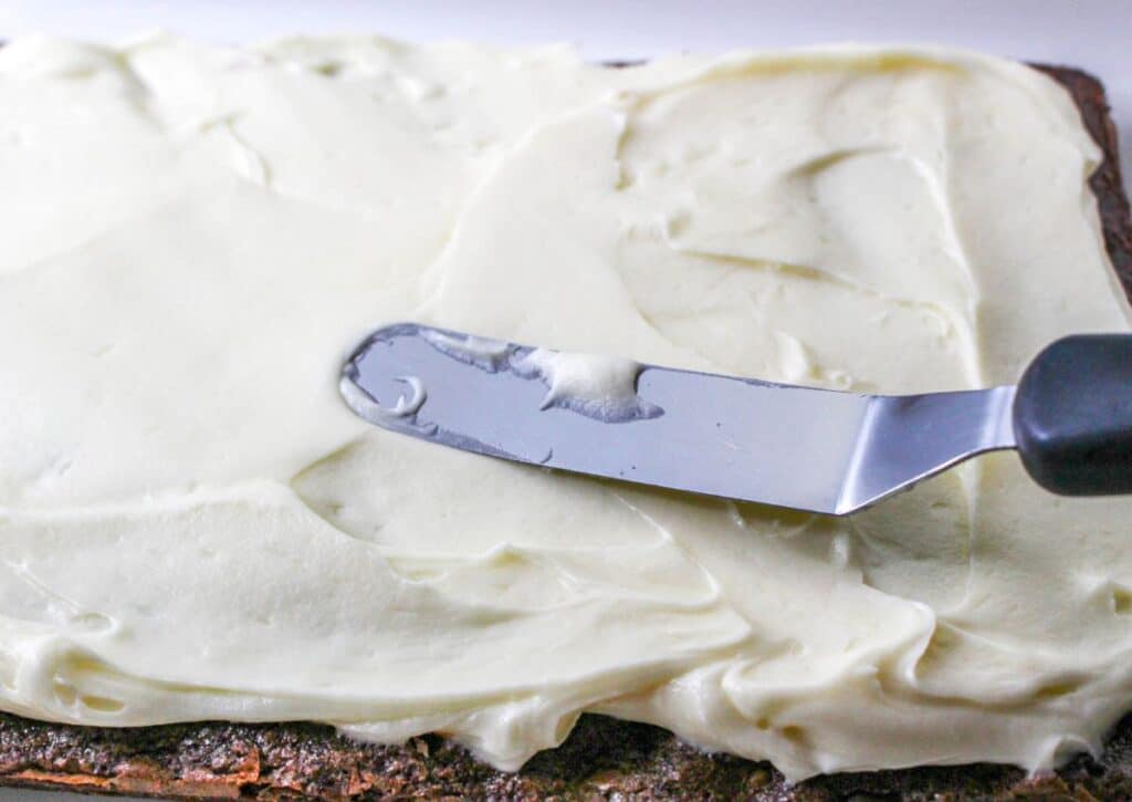 vanilla cream cheese frosting being spread on top of a rectangle of brownies