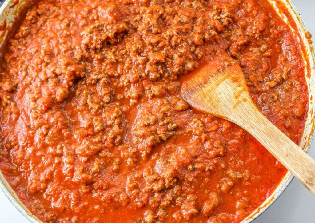 pasta sauce being mixed with cooked ground beef in a large skillet with a wooden spoon