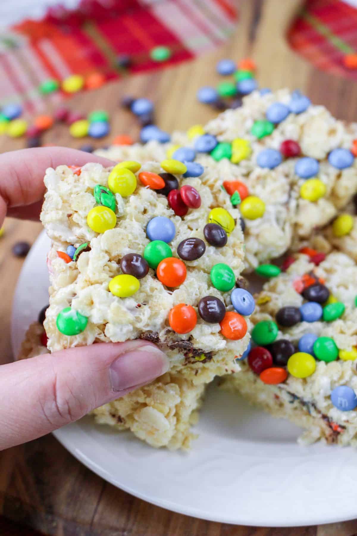 m&m rice krispie treat held in a woman's hand above a white plate stacked with the treats