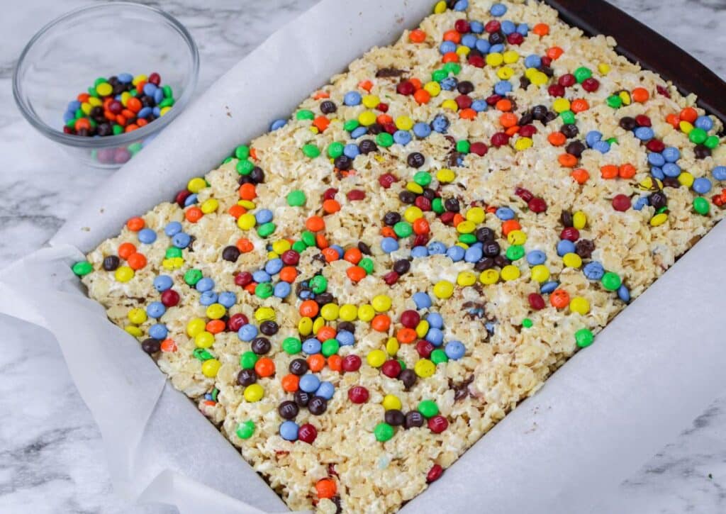 m&m's being sprinkled on top of rice krispies treats in a parchment lined baking dish