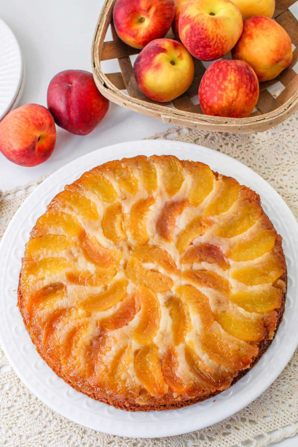 off center image of uncut peach upside down cake on a white plate. with a basket of peaches in the background