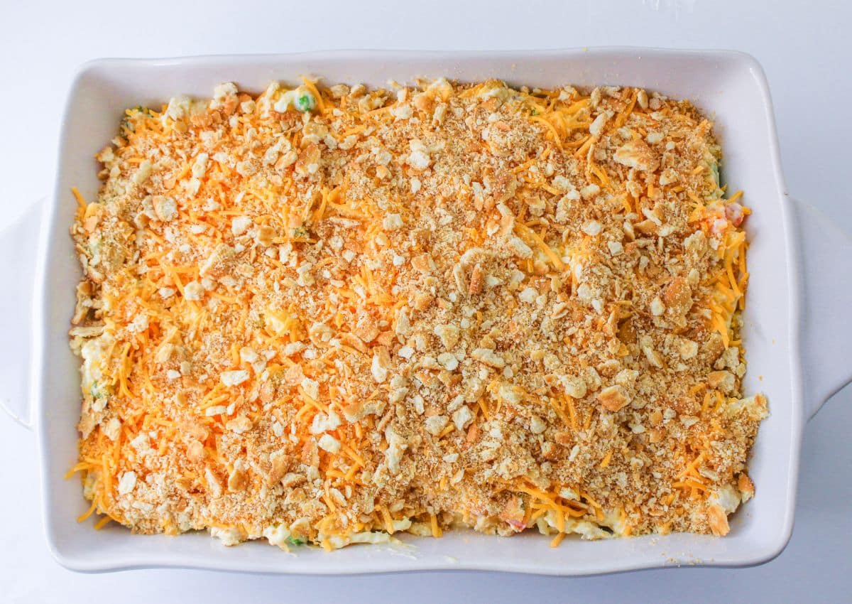 crumbled crackers and shredded cheese being sprinkled on top of an unbaked casserole.