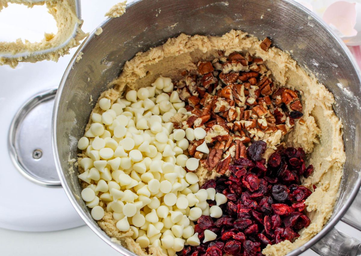 white chocolate chips, pecans, and dried cranberries being added to cookie dough in a stand mixer.