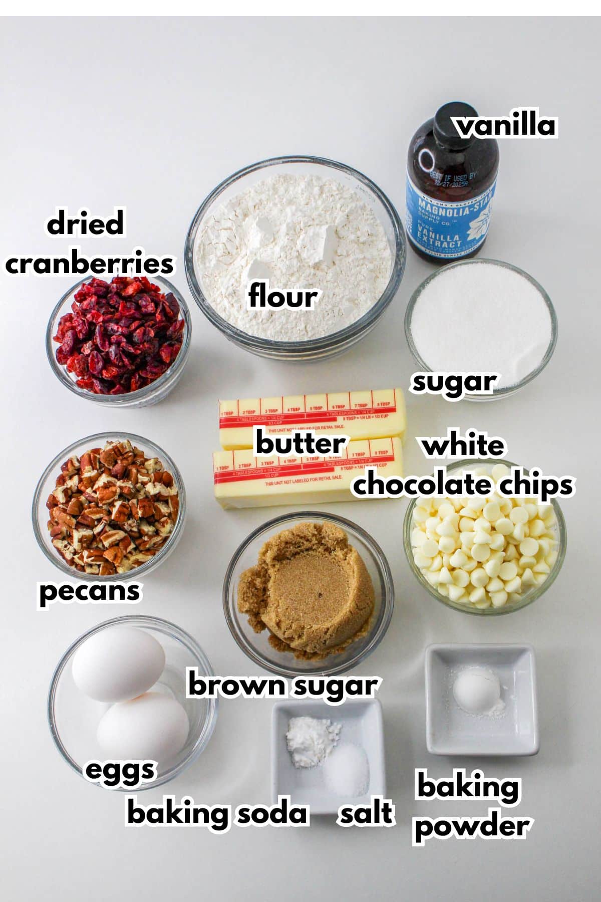 bowls of vanilla extract, flour, dried cranberries, pecans, butter, sugar, brown sugar, eggs, white chocolate chips, baking soda, baking powder, and salt.