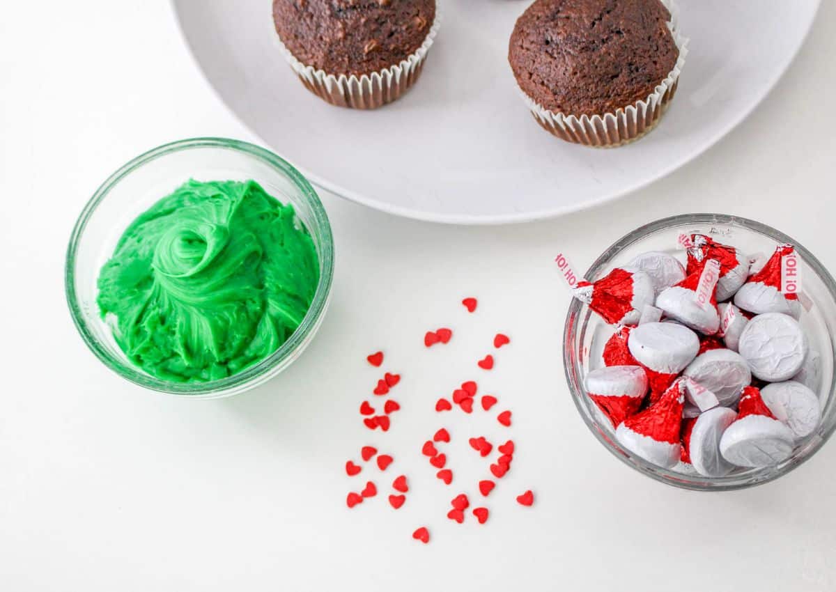 bowls of green frosting, Santa hat hershey's kisses, heart shaped sprinkles and a plate of baked chocolate cupcakes.