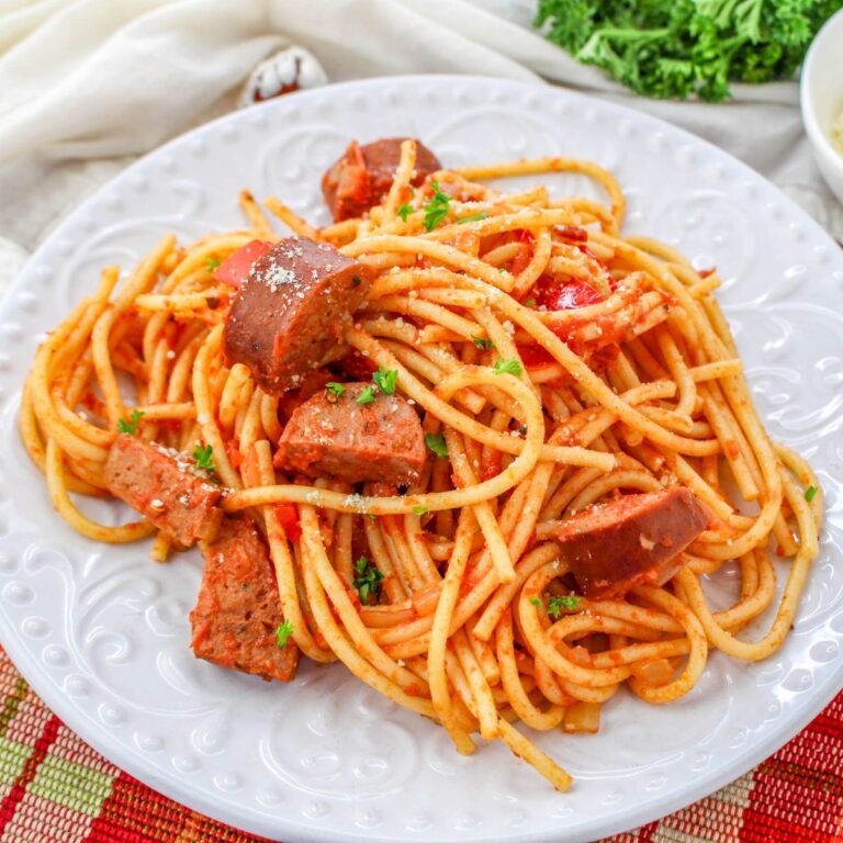 A plate of Andouille Sausage Spaghetti topped with rich tomato sauce and sliced sausage, garnished with chopped parsley, on a white decorative plate.