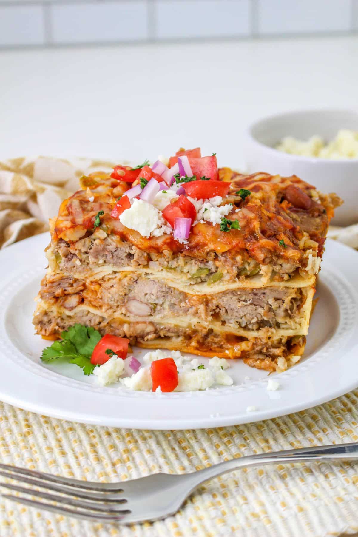 slice of pork carnitas casserole on a plate topped with queso fresco, diced onion, and diced tomatoes.