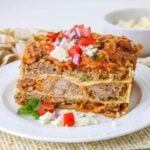 slice of pork carnitas casserole on a plate topped with queso fresco, diced onion, and diced tomatoes.