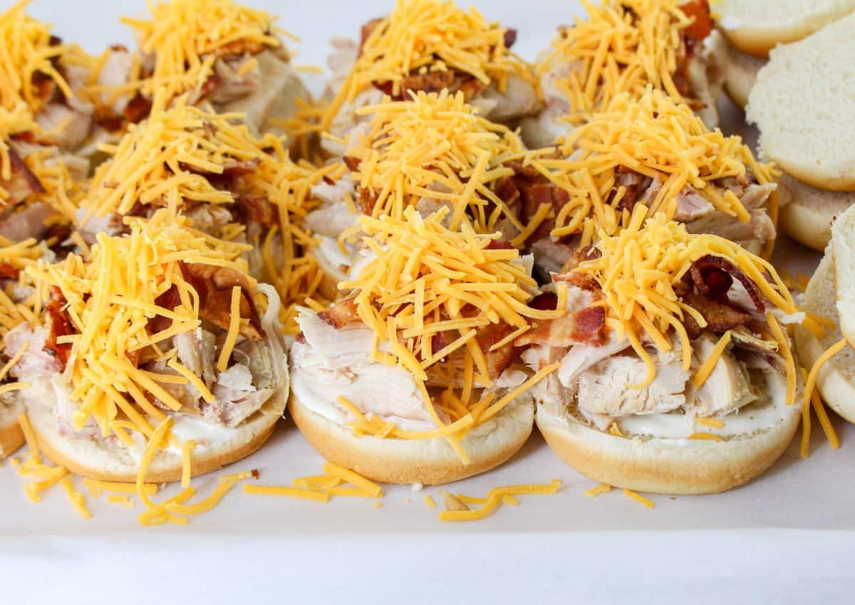 chicken, bacon, and shredded cheddar cheese being layered on top of buns.