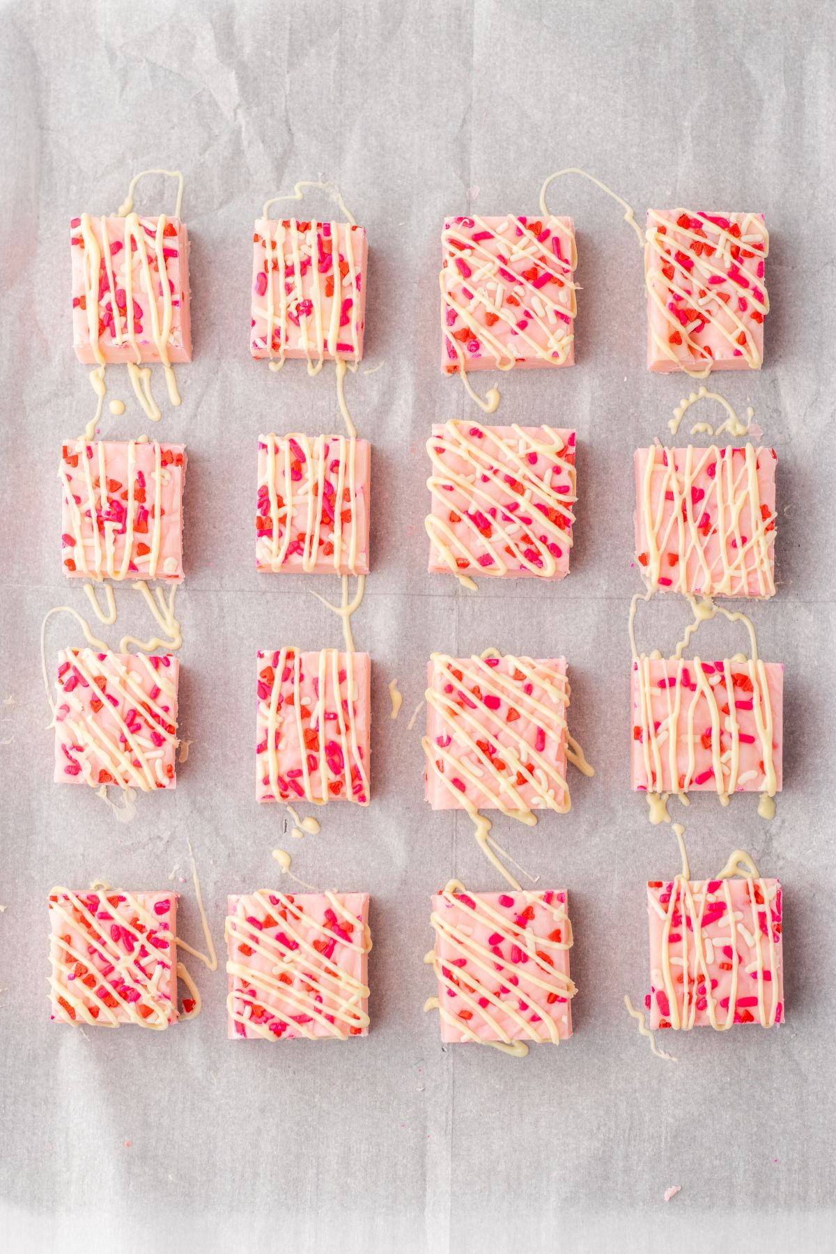 pieces of pink fudge cut into squares on a parchment lined baking sheet being drizzled with white chocolate.