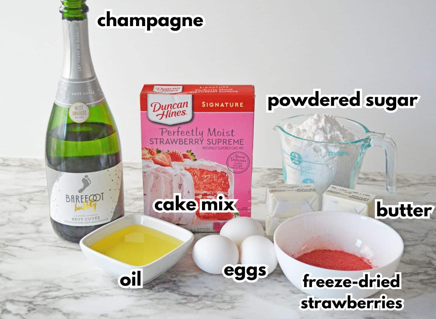 champagne, powdered sugar, oil, cake mix, eggs, butter, and freeze-dried strawberries on a backdrop.