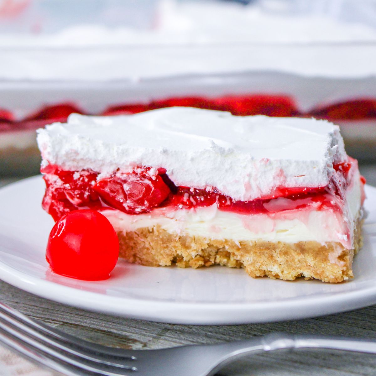 A slice of cherry cheesecake lush dessert with whipped topping on a plate, garnished with a maraschino cherry.