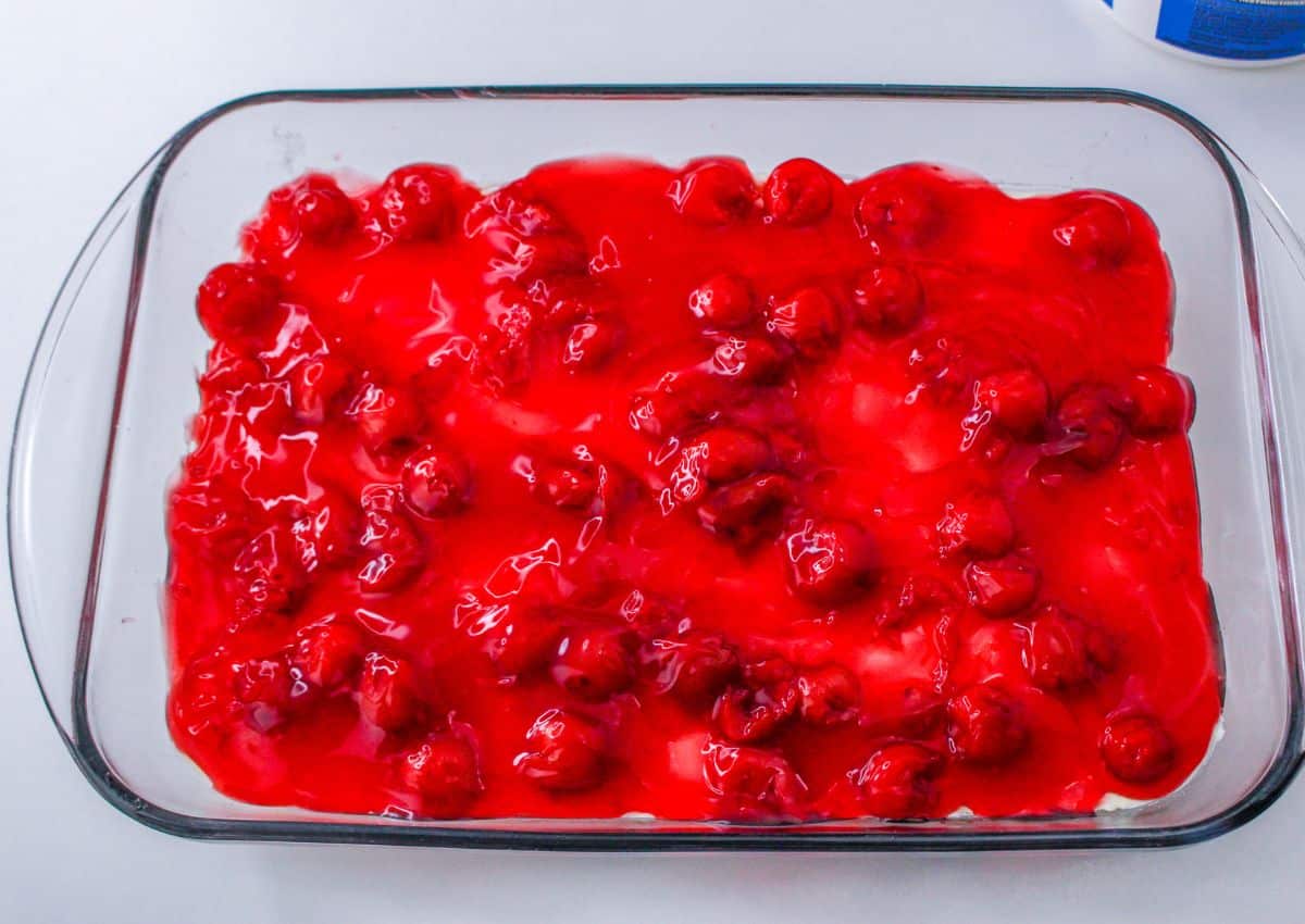 cherry pie filling being spread on top of a dessert in a glass casserole dish.