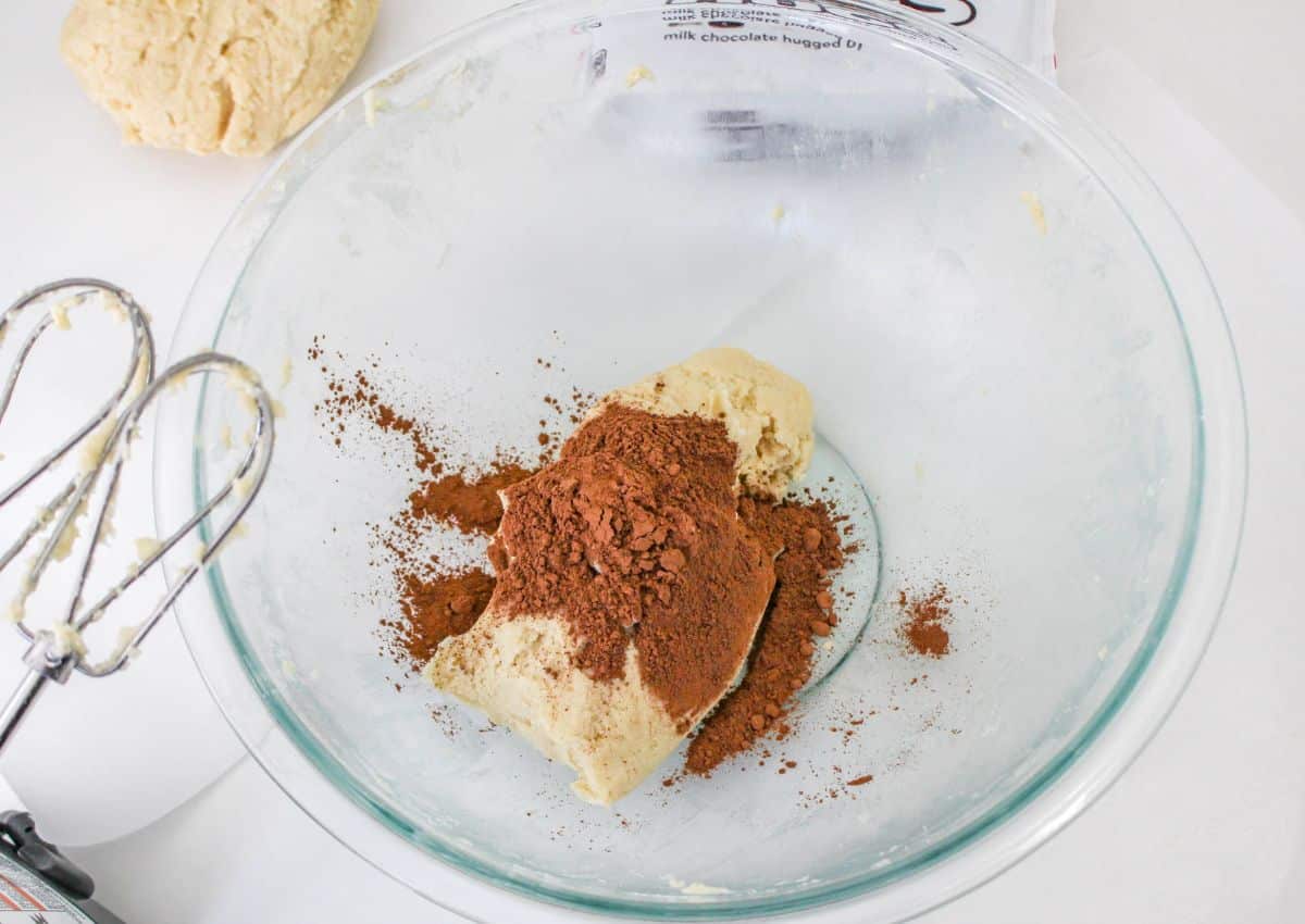 cocoa powder being added to cookie dough in a glass mixing bowl.