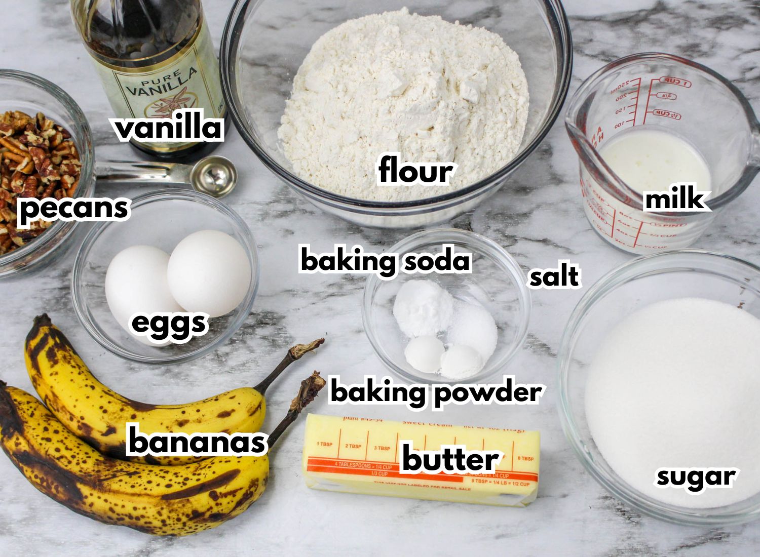 bowls of pecans, vanilla extract, flour, milk, eggs, baking soda, baking powder, salt, sugar, butter and bananas on a counter.