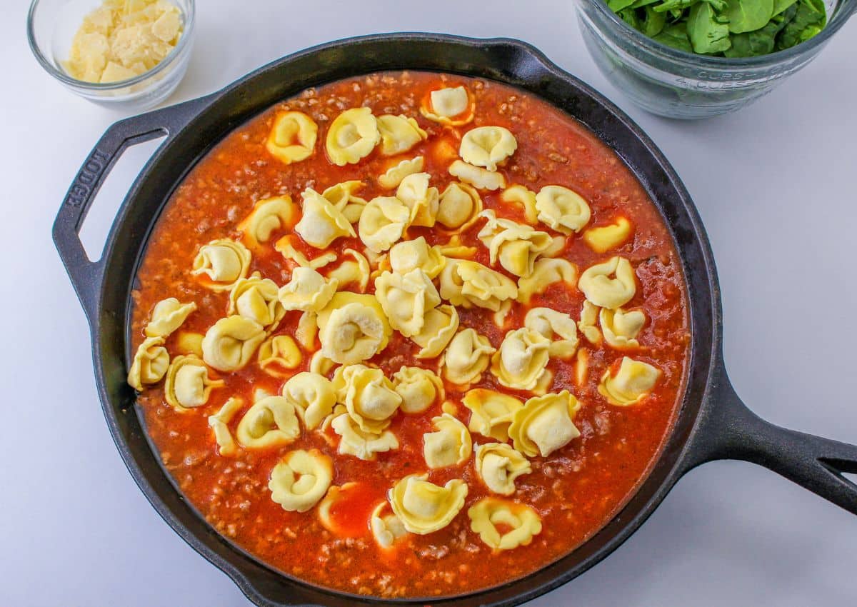raw tortellini being added to a cast iron skillet full of tomato sauce and broth.