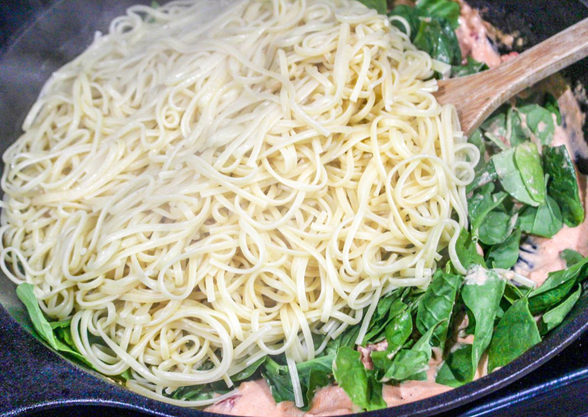 linguine and spinach being added to a cast iron skillet.