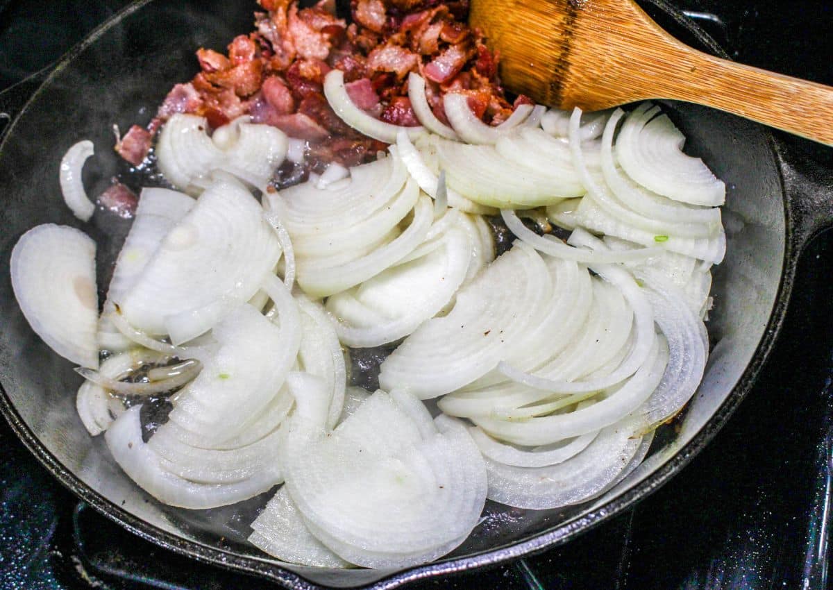 onions being cooked in a cast iron skillet with chopped bacon.