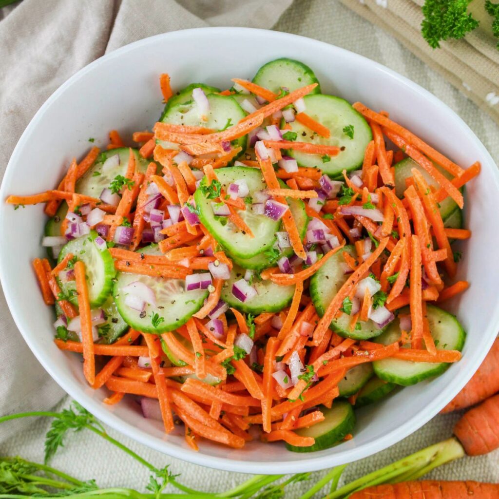 A white bowl filled with cucumber carrot salad featuring sliced cucumbers, shredded carrots, and chopped red onions, garnished with parsley.