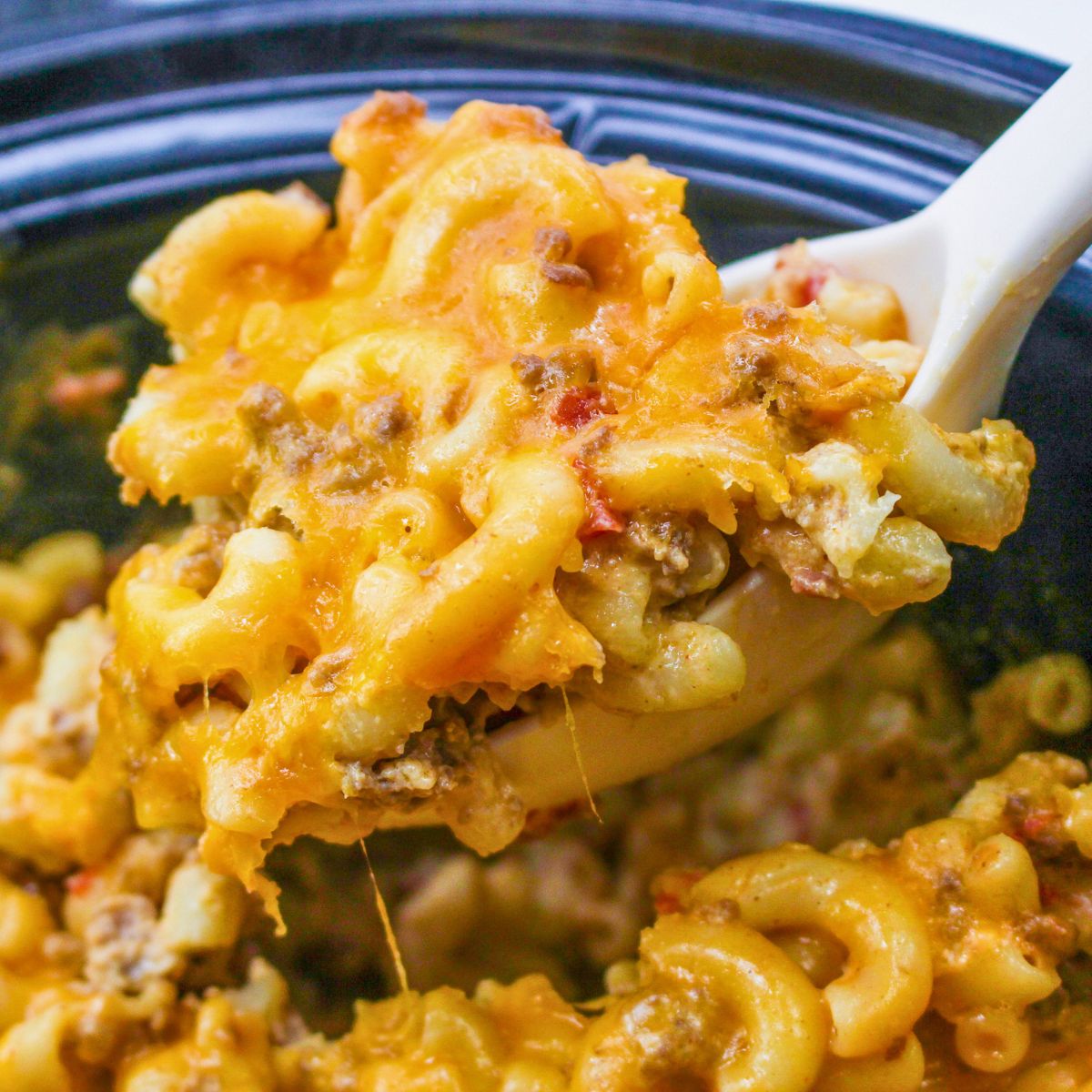A close-up of a spoon scooping cheesy slow cooker taco mac and cheese, mixed with ground beef and tomato, from a black pot.