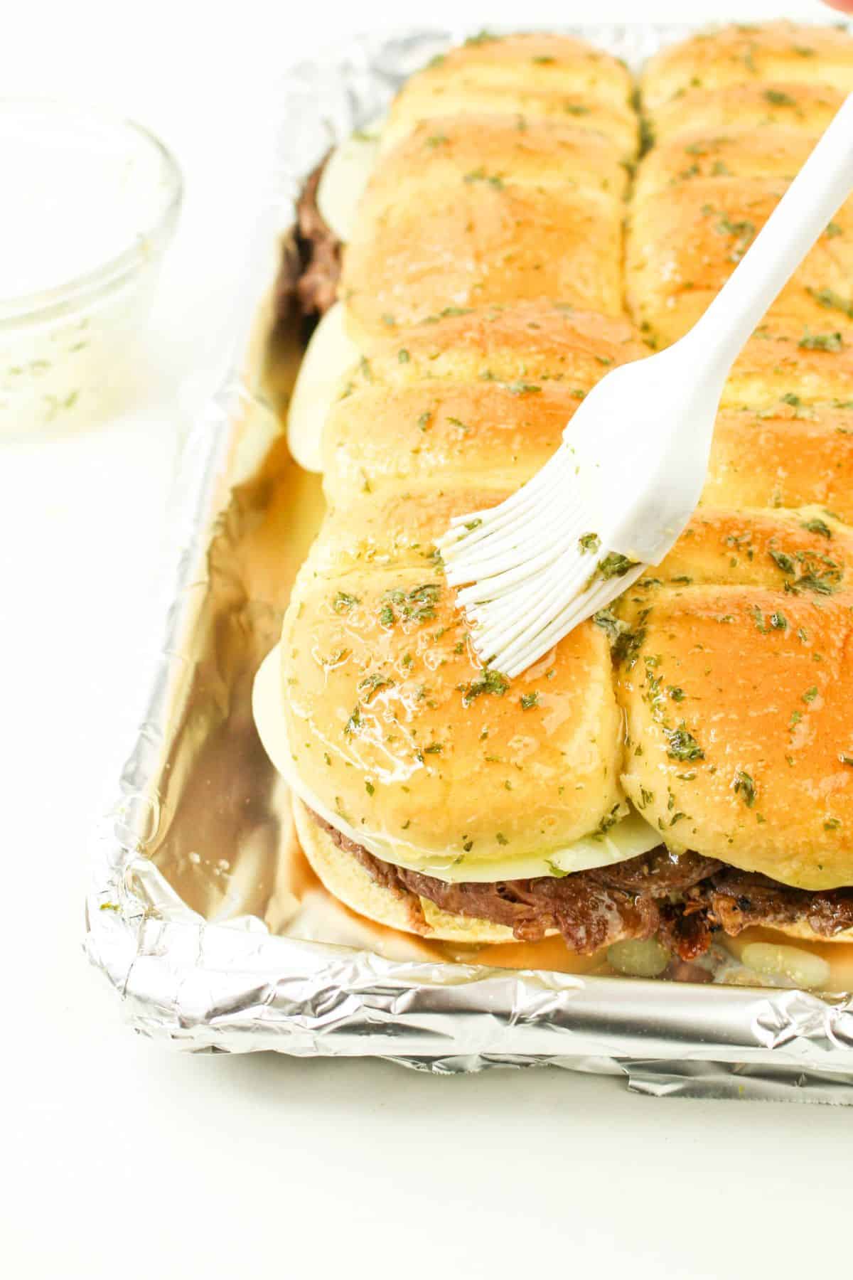 garlic butter being brushed on top of sliders on a baking sheet.