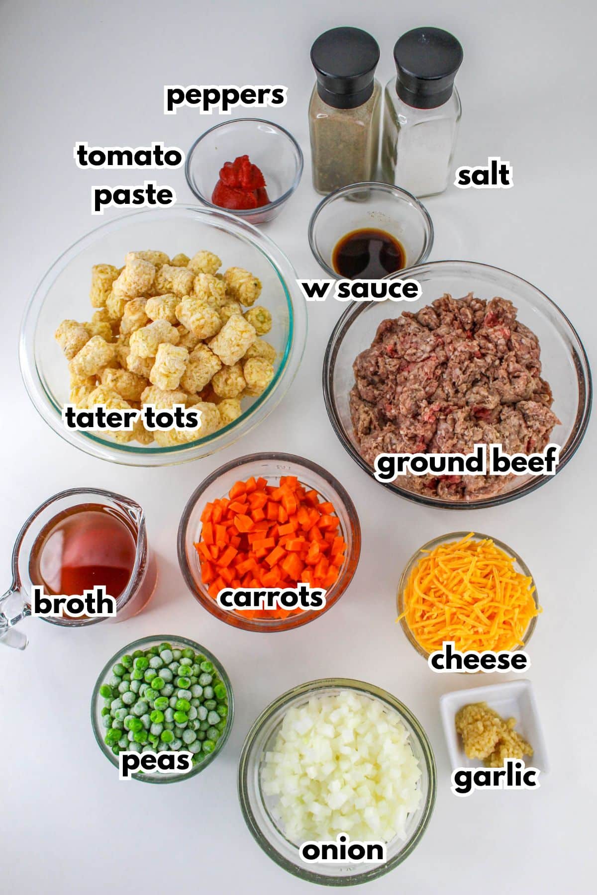 bowls of pepper, salt, tomato paste, w sauce, tater tots, ground beef, broth, carrots, peas, onion, cheese and garlic on the counter.