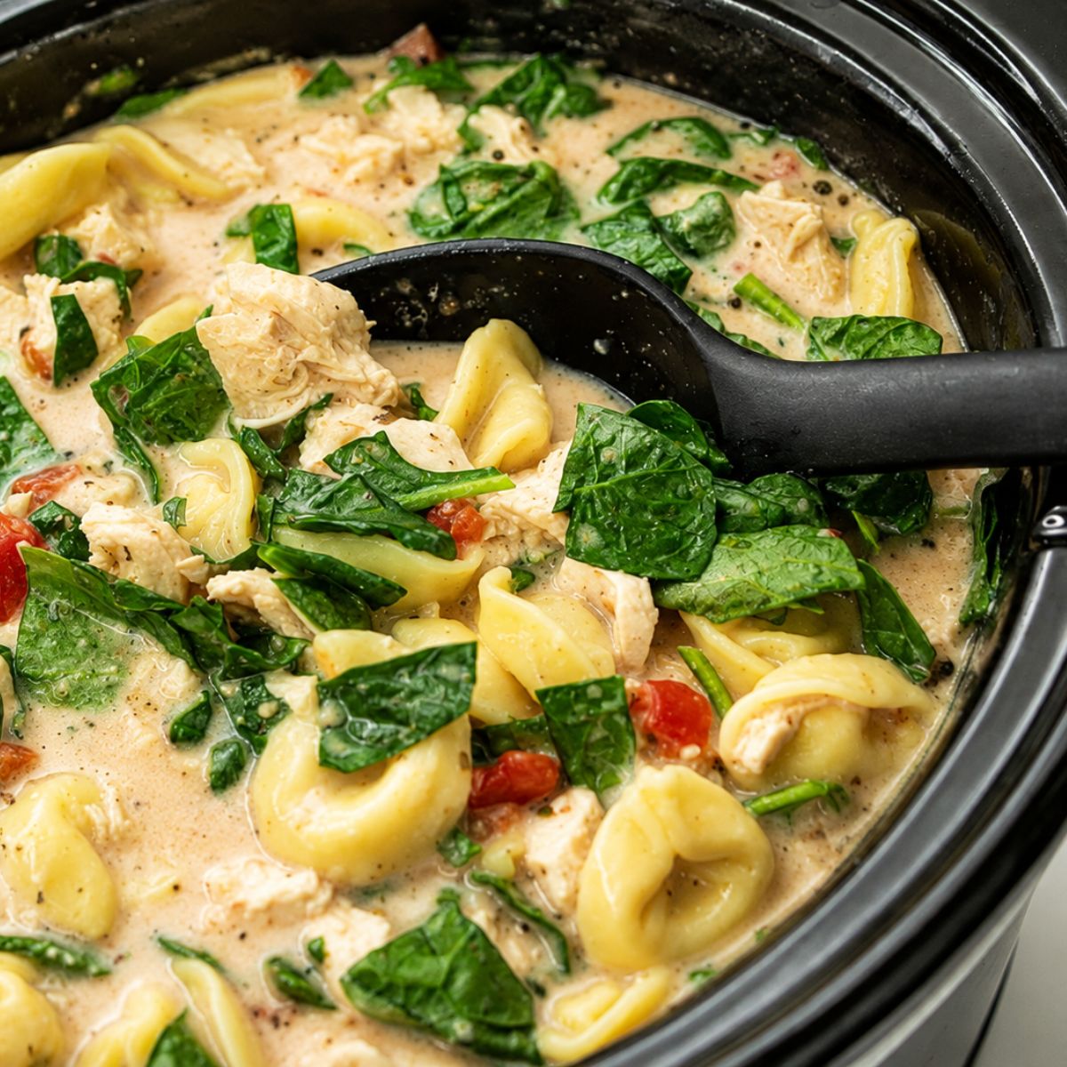 A slow cooker filled with creamy slow cooker chicken and tortellini soup, featuring tender spinach, juicy chicken, diced tomatoes, and a black serving spoon.