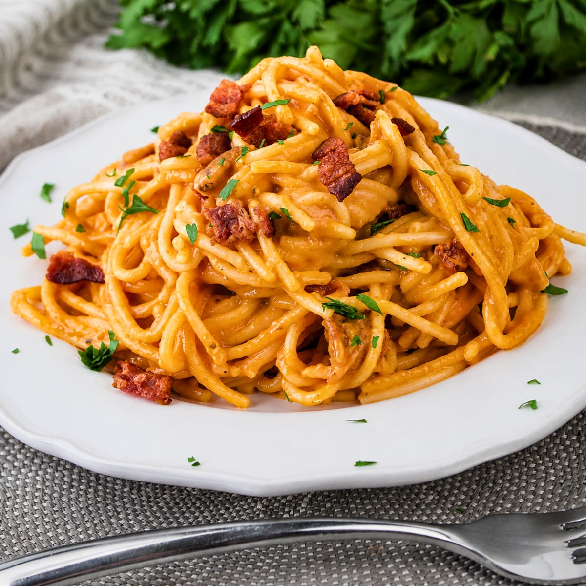 A plate of Pumpkin Carbonara, featuring spaghetti coated in a creamy orange sauce, is topped with crumbled bacon and chopped parsley and sits on a white plate next to a fork.
