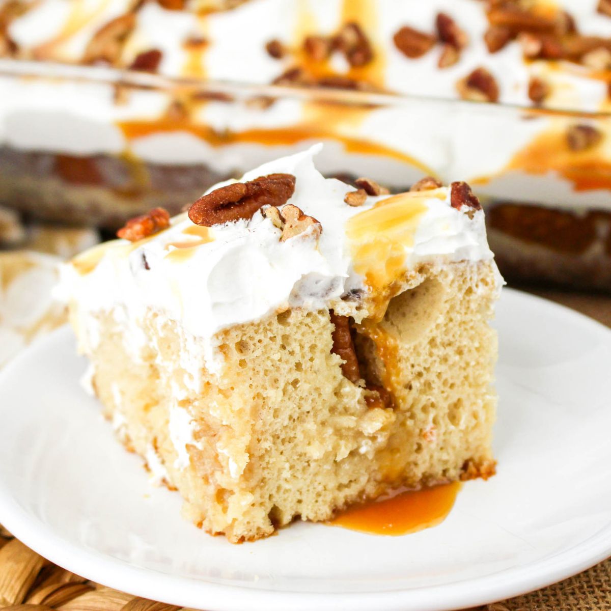 A slice of Butter Pecan Poke Cake topped with whipped cream, caramel sauce, and pecans sits on a white plate, with the rest of the cake in the background.