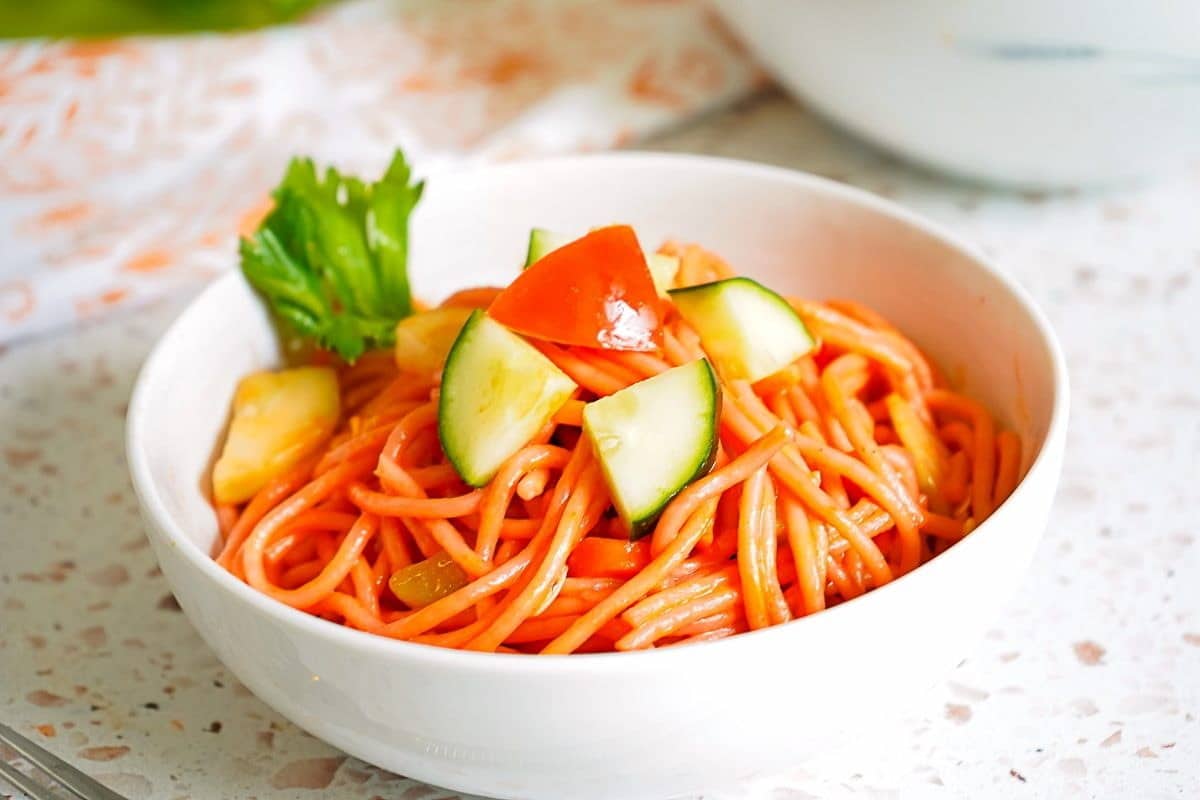 A bowl of spaghetti topped with chunks of cucumber, tomato, and pieces of lettuce on a speckled countertop.