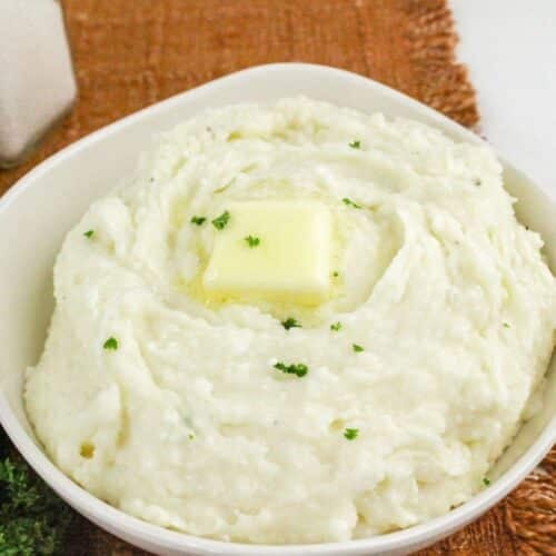 A bowl of mashed potatoes with a pat of butter on top, garnished with parsley, set on a woven placemat.