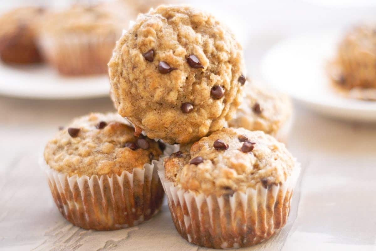 A stack of four chocolate chip muffins on a light surface, with one muffin resting on top of the others.