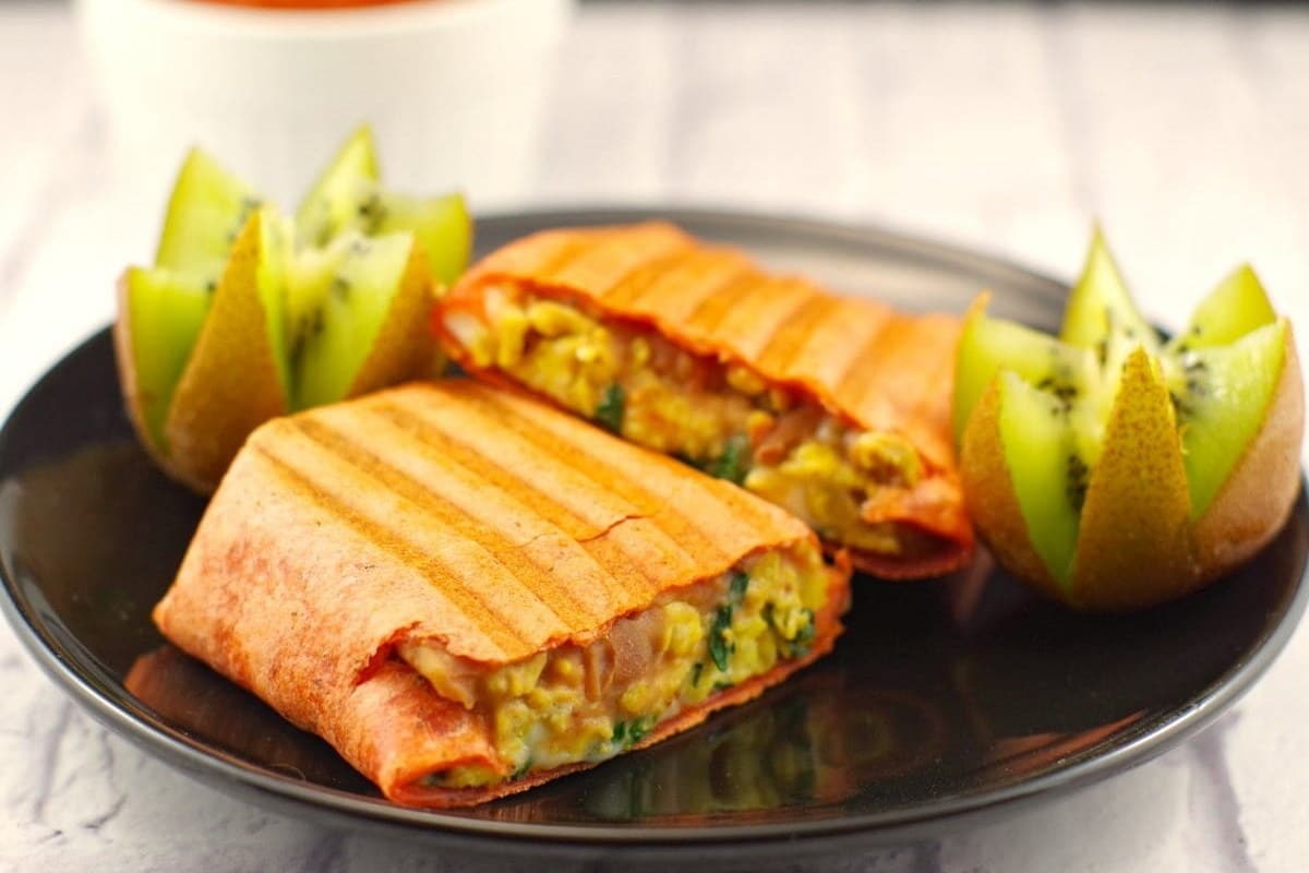 Grilled wrap with cheese and vegetables on a black plate, accompanied by sliced kiwi.