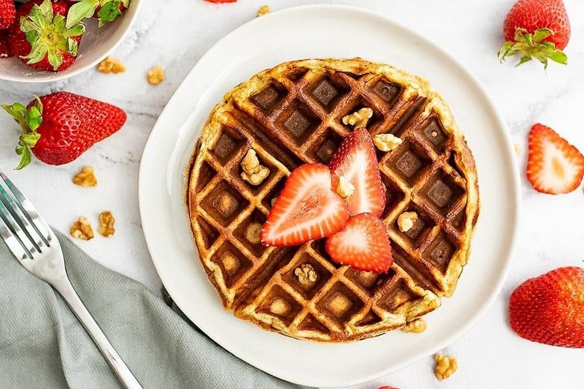 A waffle topped with sliced strawberries and walnuts on a white plate. Nearby are fresh strawberries, walnuts, a fork, and a gray napkin.