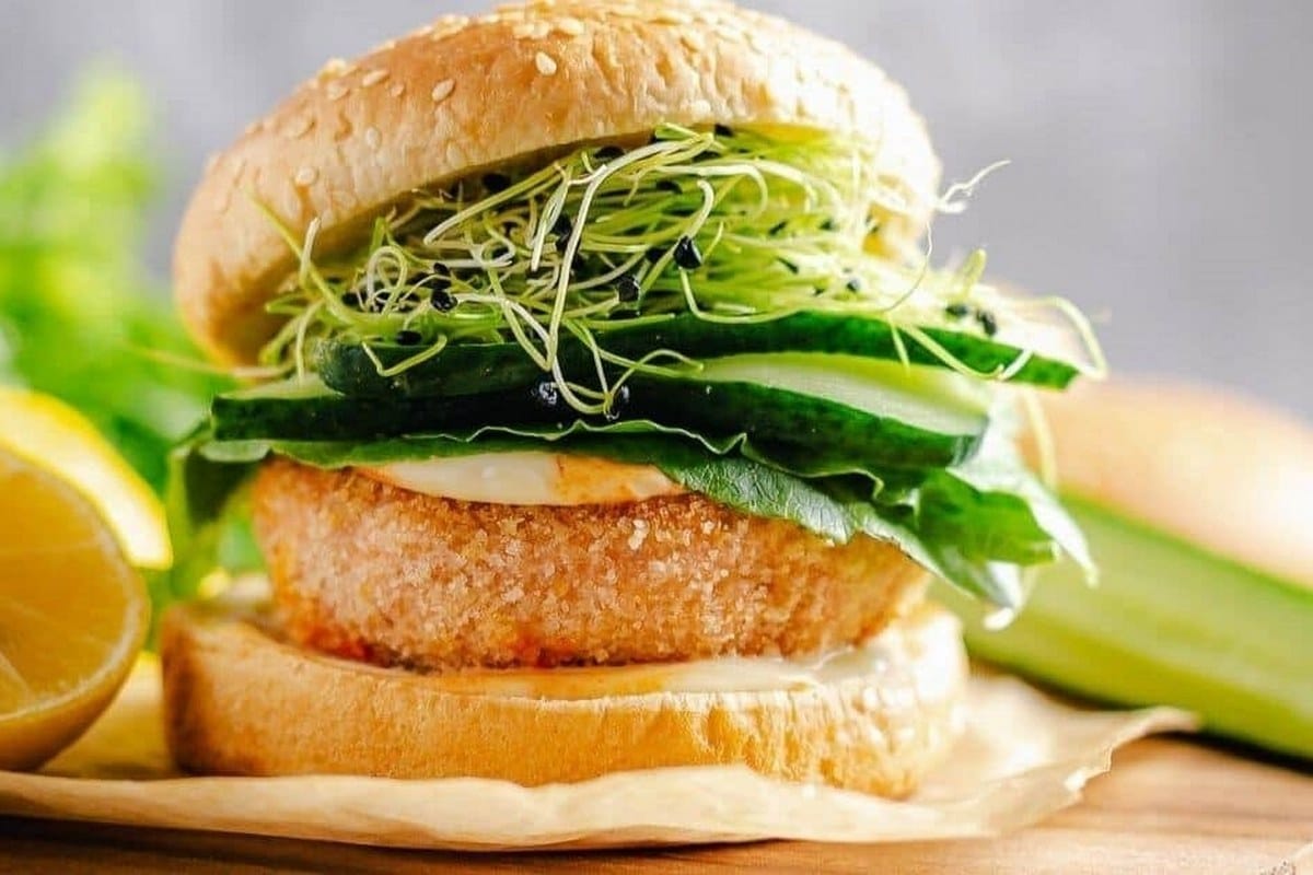 A veggie burger with lettuce, cucumber slices, and alfalfa sprouts on a sesame seed bun, placed on parchment paper. A lemon half and cucumber are in the background.