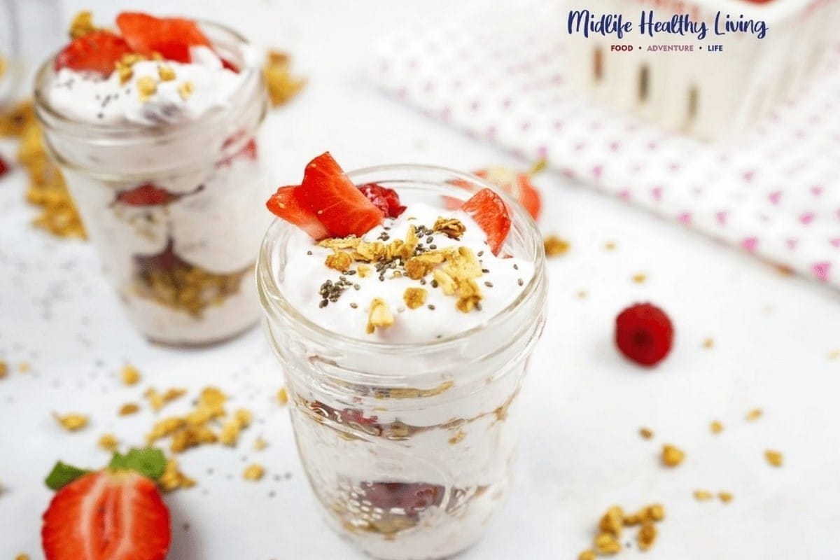 Two jars filled with layered yogurt, granola, and strawberries on a white surface.