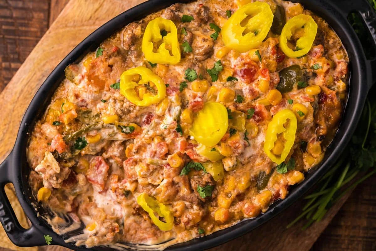 A baked dish in a black casserole, topped with yellow pepper slices and a creamy mix containing corn, tomatoes, and herbs.