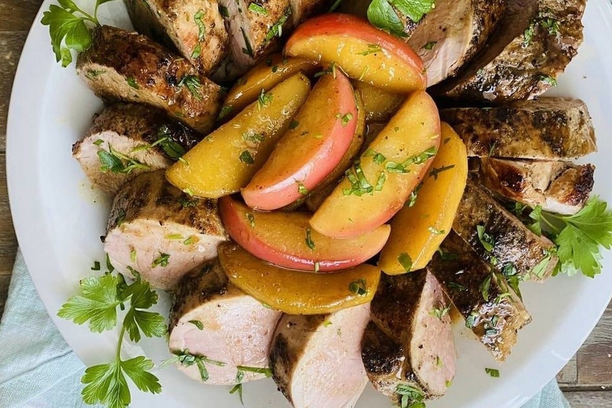 Sliced roast pork tenderloin topped with apple slices and herbs on a white plate, garnished with parsley.