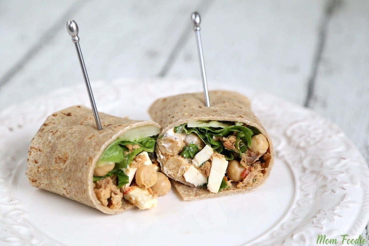 A whole wheat wrap cut in half, filled with chickpeas, spinach, feta cheese, and various ingredients, secured with skewers, placed on a white plate.