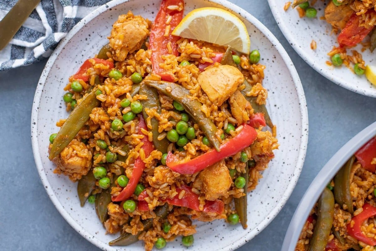 A plate of rice with chicken, red peppers, peas, and snow peas, garnished with a lemon wedge.