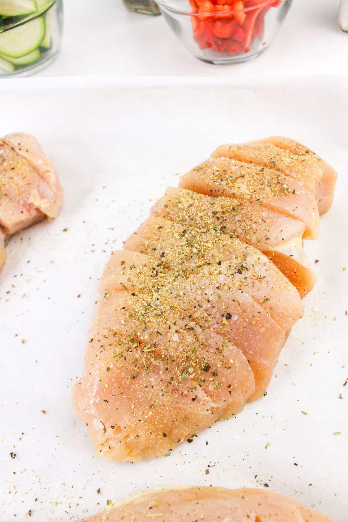 Seasoned raw chicken breast slices on a white tray, prepped with herbs and spices.