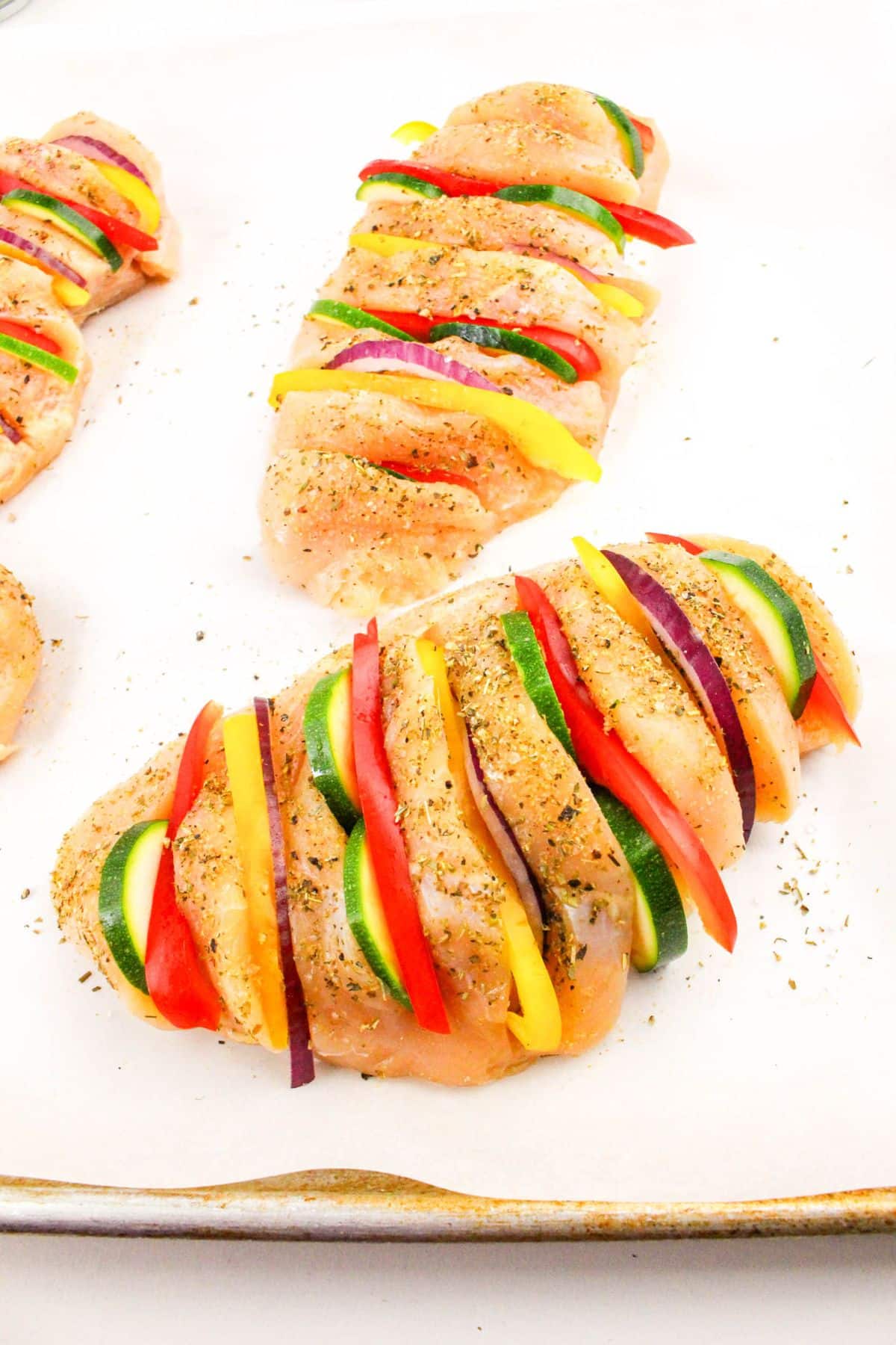 Raw chicken breasts with incisions, stuffed with slices of zucchini, red onion, and bell peppers, seasoned on parchment paper.