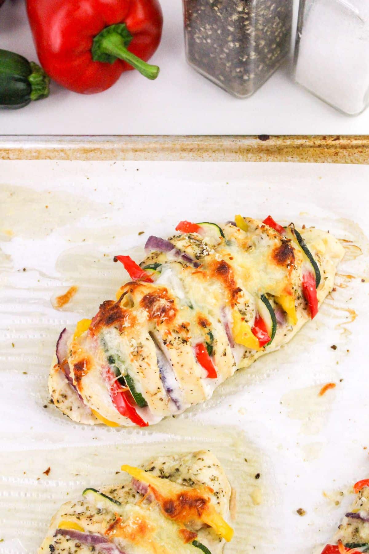 Sliced baked chicken breast stuffed with red peppers, yellow peppers, zucchini, and red onions, topped with melted cheese on a baking sheet. Salt and pepper shakers in the background.