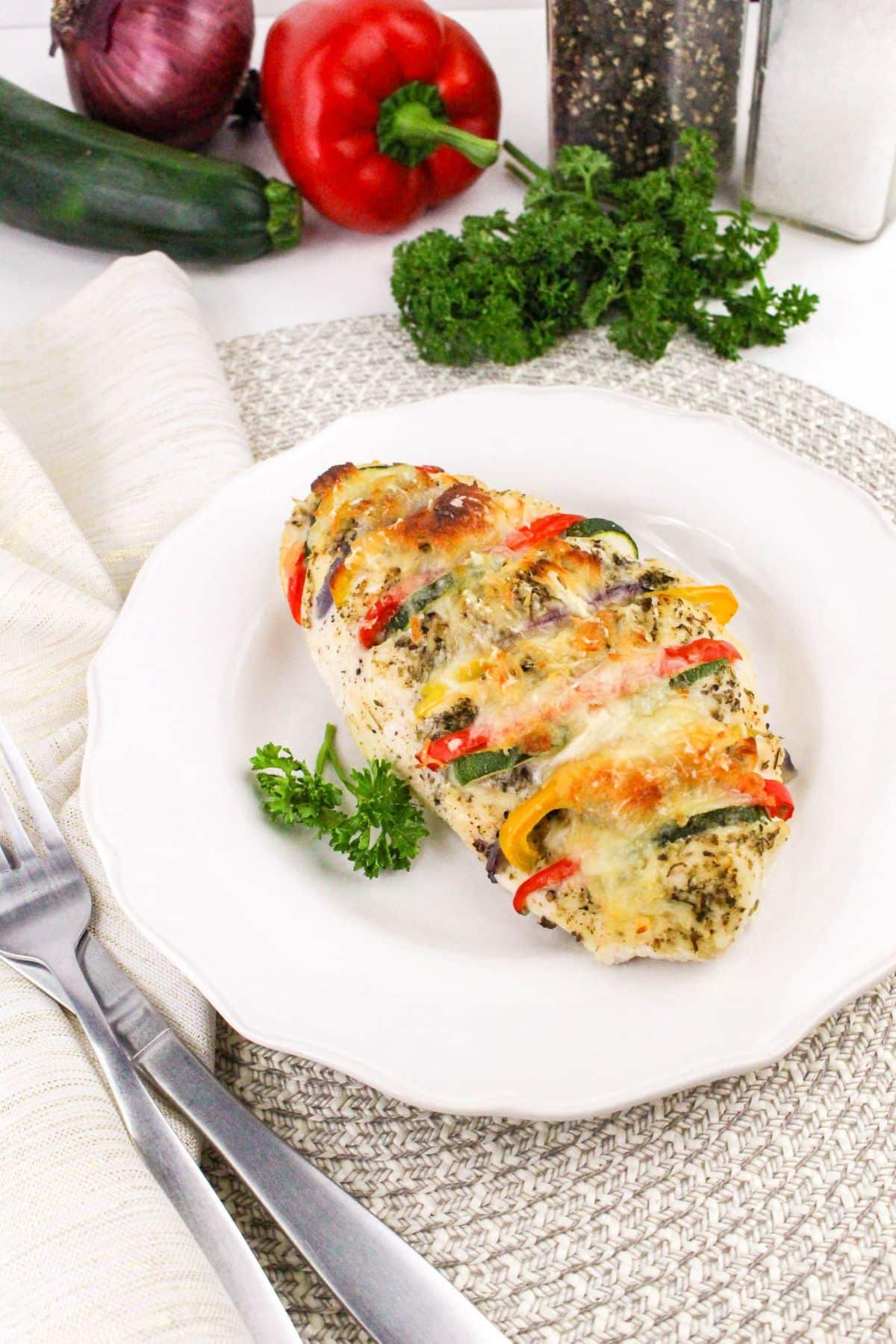 A stuffed chicken breast with colorful peppers and melted cheese on a white plate. A fork and knife are beside it, with vegetables and herbs in the background.