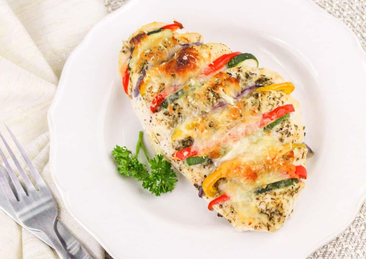 Baked chicken breast stuffed with slices of zucchini, bell peppers, and cheese on a white plate, garnished with parsley.